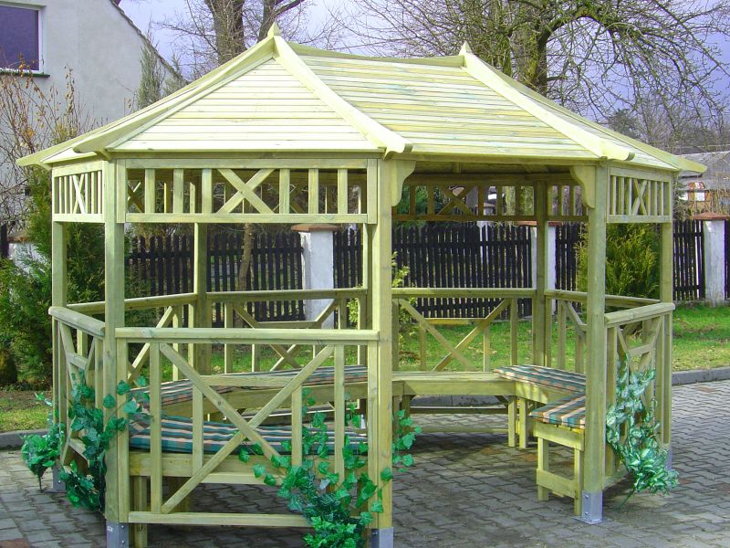 Sześcioboczna, drewniana altana ogrodowa z siedziskami, ozdobiona sztucznym bluszczem, na tle trawnika i ogrodzenia.